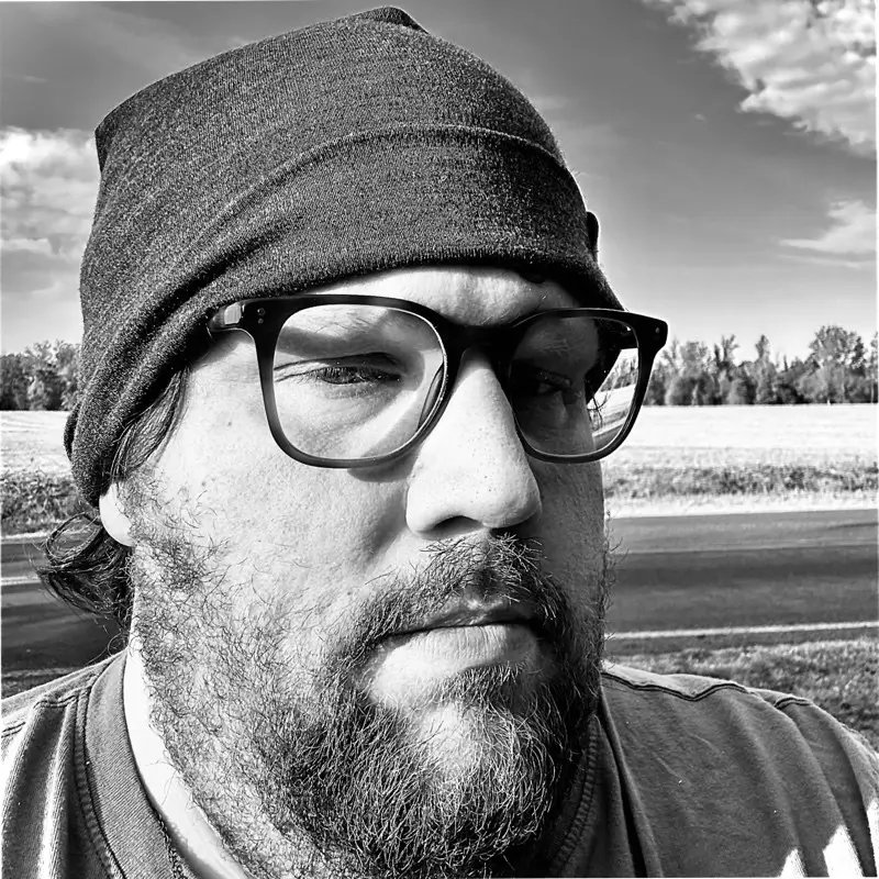 A black and white photo of Josh in front of a field. It is wearing a wool beanie and glasses.