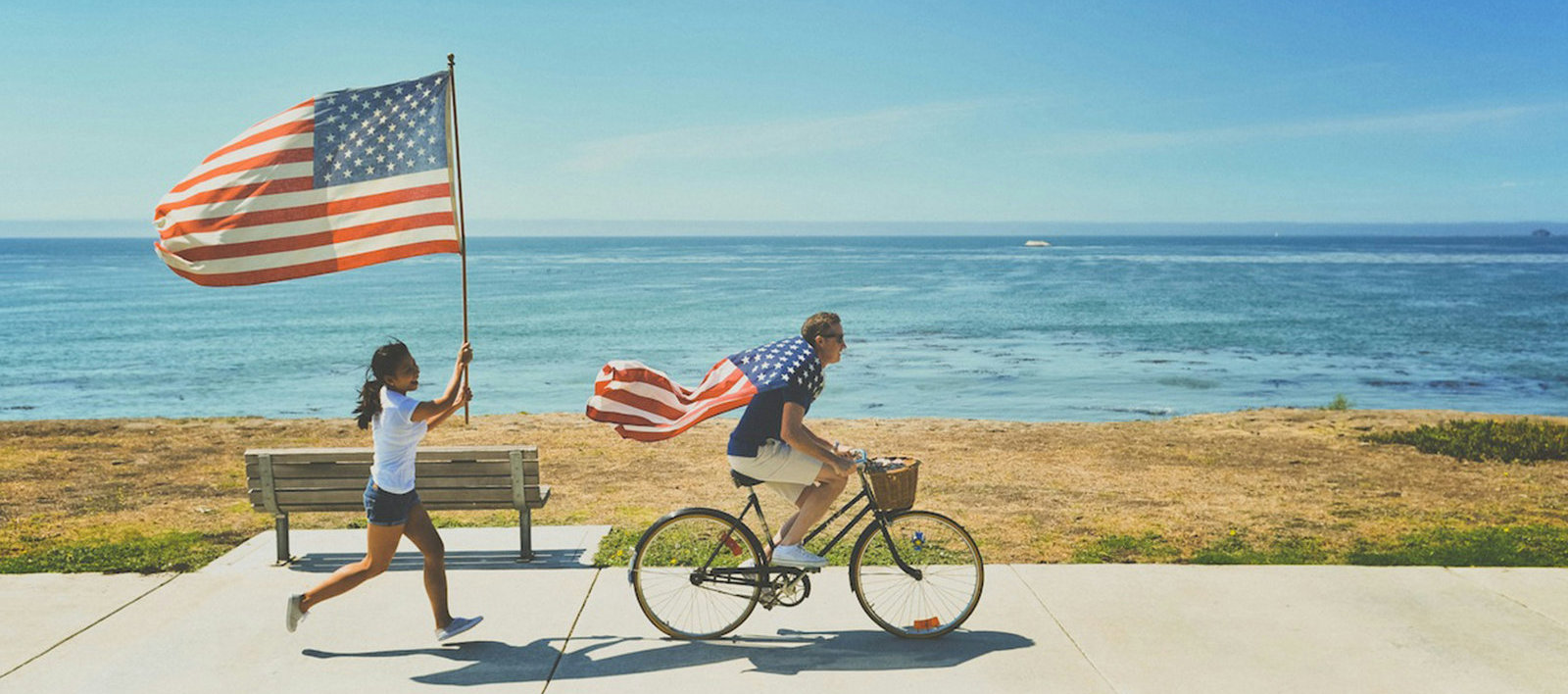 A person rides a bike on a path next to an ocean wearing an American flag as a cape. Behind them a girl follows running carrying a large American flag.